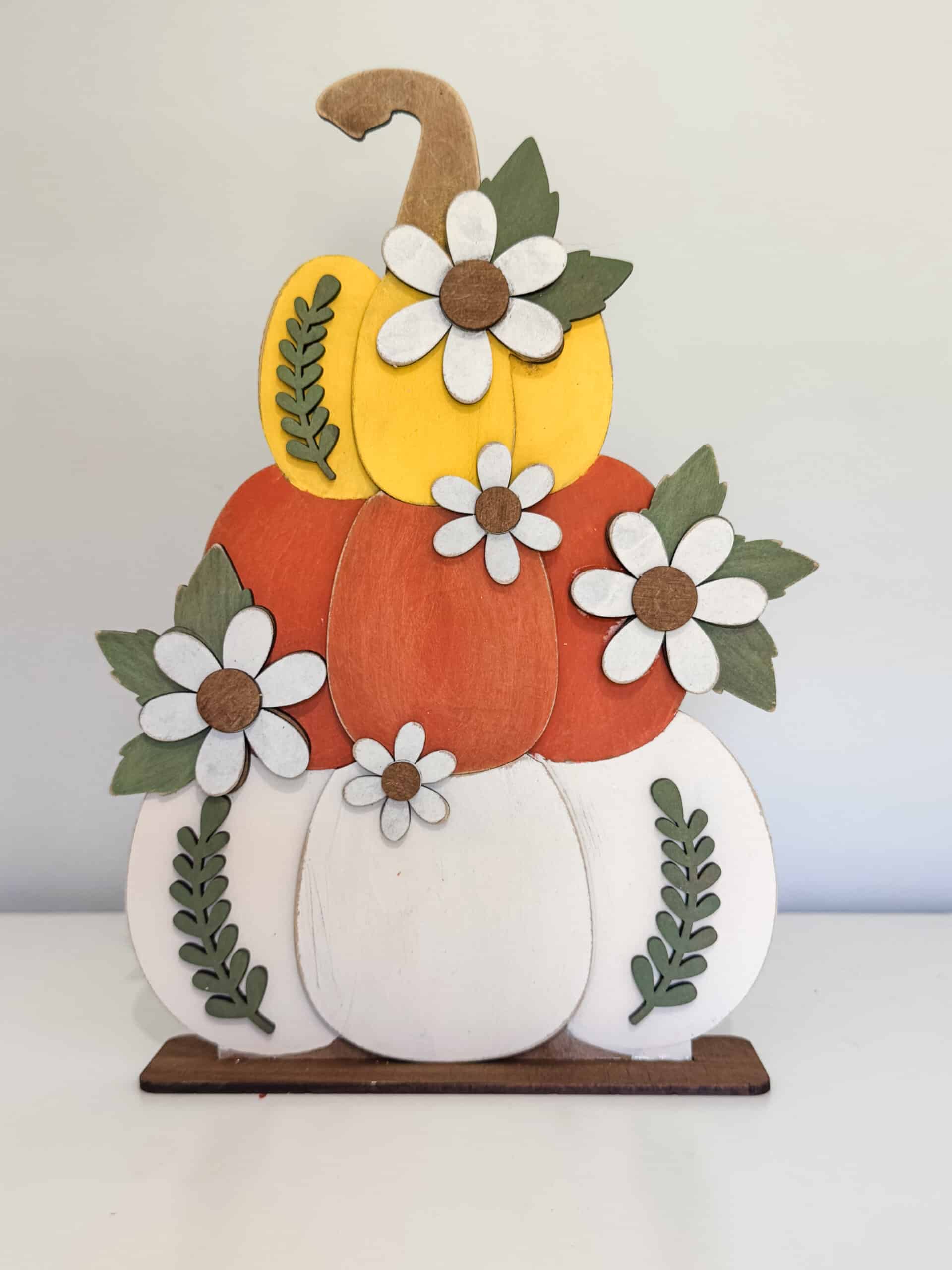 Finished wooden pumpkin stack with a yellow top, orange middle, cream bottom, white flowers, green leaves, and a brown stand.