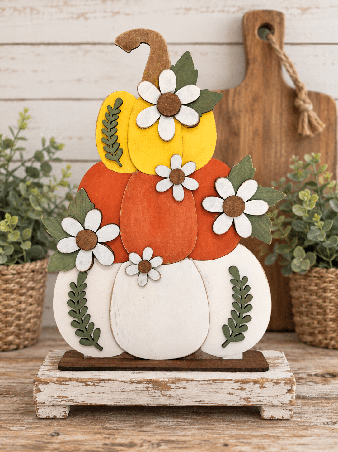 Yellow, orange, and cream wooden pumpkin stack with white flowers and green leaves, staged on a rustic riser beside greenery.