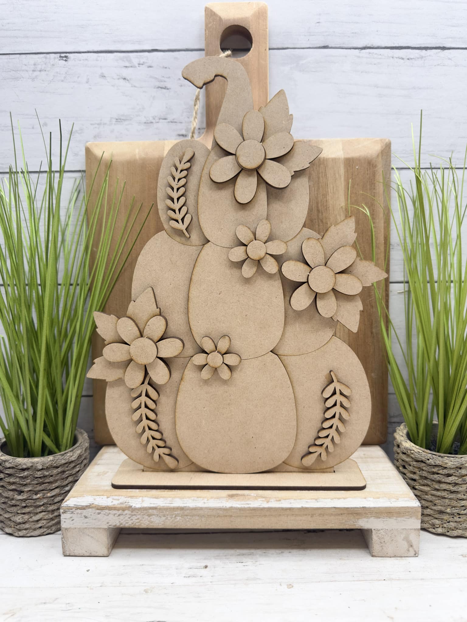 Unpainted floral stacked pumpkin shelf sitter displayed on a distressed white riser between potted greenery.