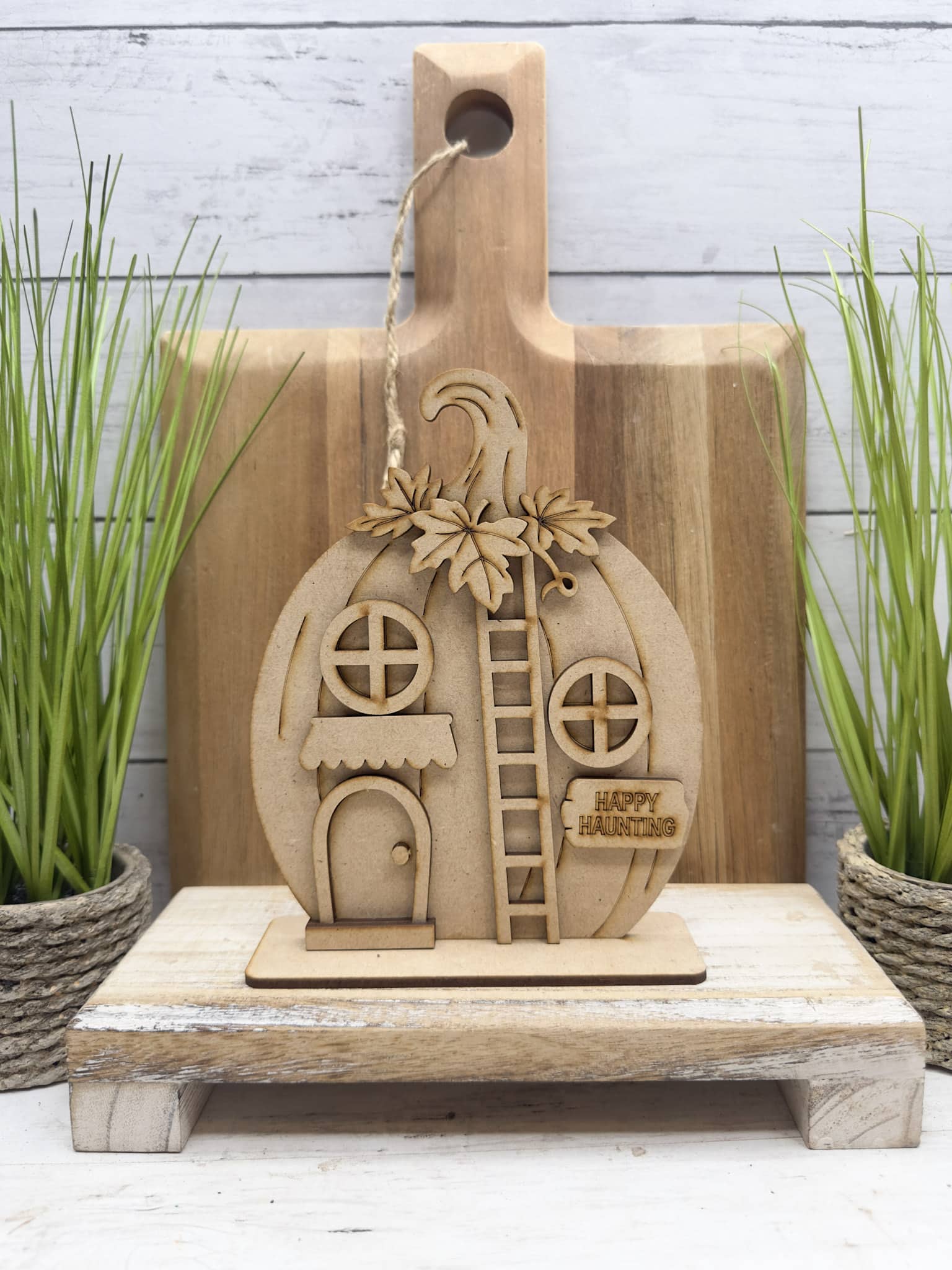 Unfinished Happy Haunting Pumpkin House DIY craft kit assembled and staged on a rustic wood riser with greenery and cutting board backdrop.