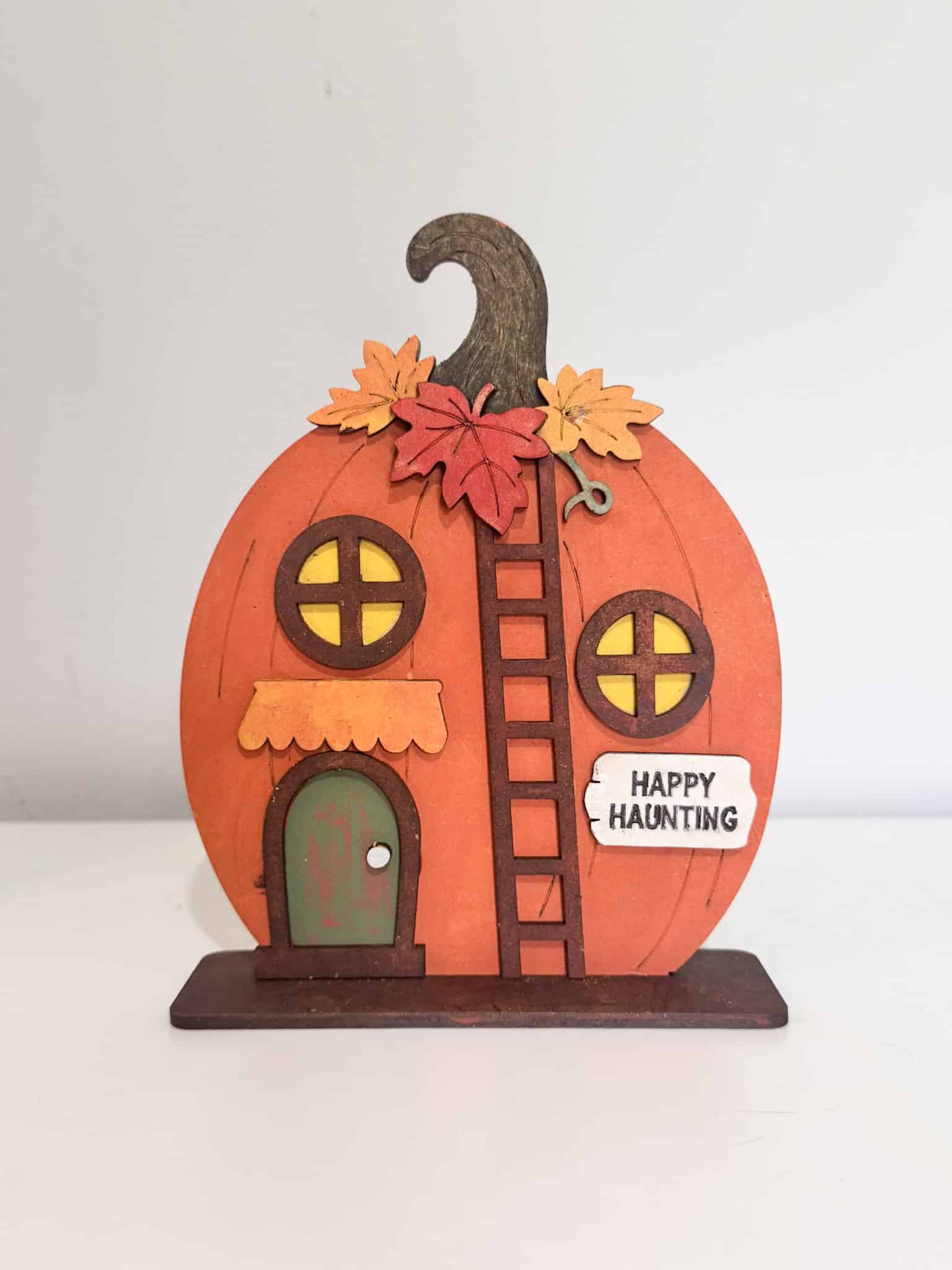 Finished Happy Haunting Pumpkin House DIY kit painted orange with fall leaves, yellow windows, distressed green door and brown ladder.