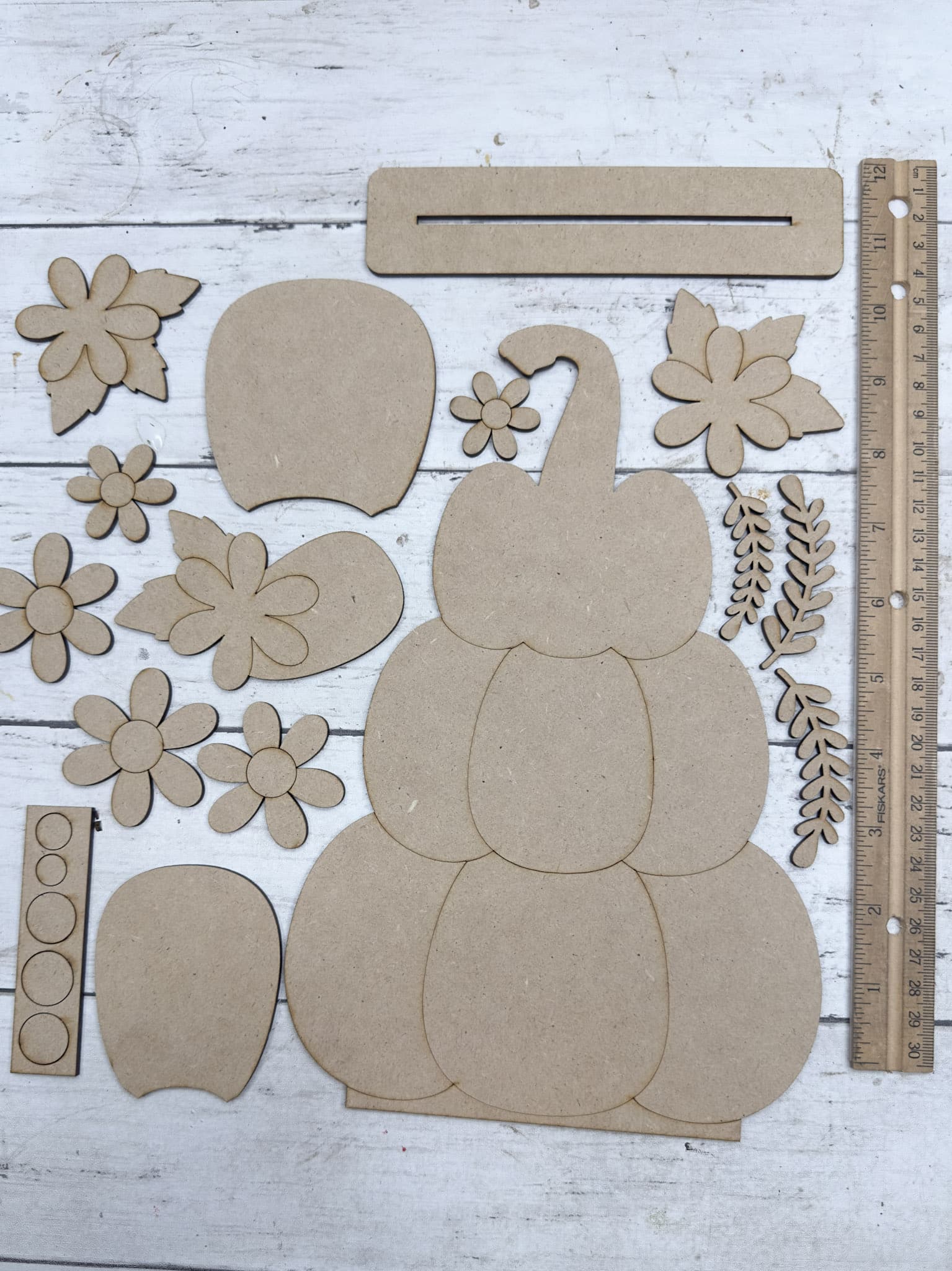 Unfinished wooden pumpkin craft kit pieces laid flat, including a stacked pumpkin backing, flowers, leaves, center layers, and stand.